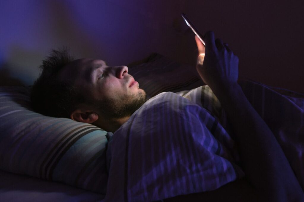布団の中で横になりながらスマホを見て眠れずにいる人物。考えごとが止まらず目が冴えている様子